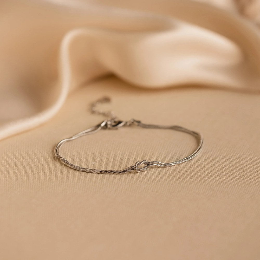 The Pomence Forever Knot Bracelet in cool steel-silver tone photographed alone, clasped into a continuous ring on warm tan satin under directional light