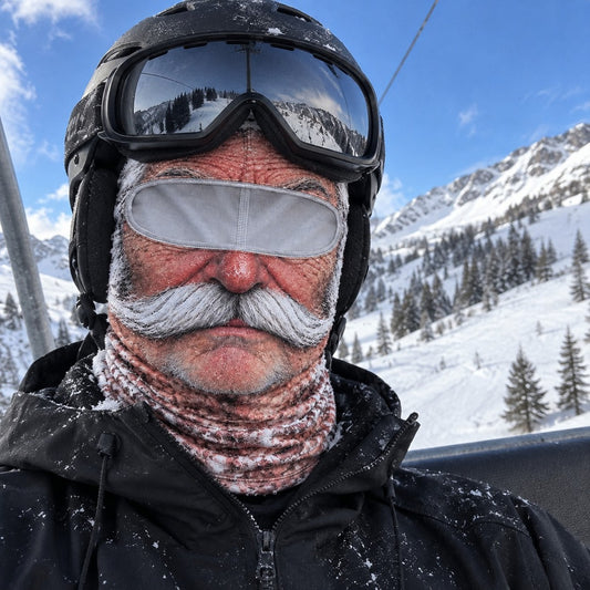 A skier on a chairlift in the Grandpa mask with ski goggles and a helmet over it, open blue sky above, snowy run below.
