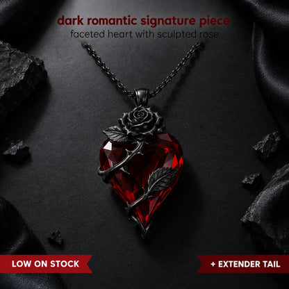 Pomence Blood Rose Necklace flat lay on black, a faceted blood red crystal heart pendant with a sculpted dark gunmetal rose and a thorned stem with two leaves on a fine gunmetal cable chain