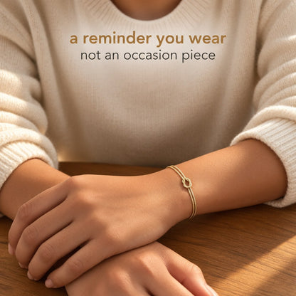 A bare wrist wearing the Pomence Forever Knot Bracelet in warm 18k-gold tone under a pushed-up sweater sleeve
