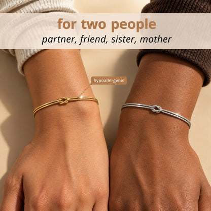 Two bare wrists side by side on a warm backdrop, one wearing the Pomence Forever Knot Bracelet in warm 18k-gold tone and one in cool steel-silver tone