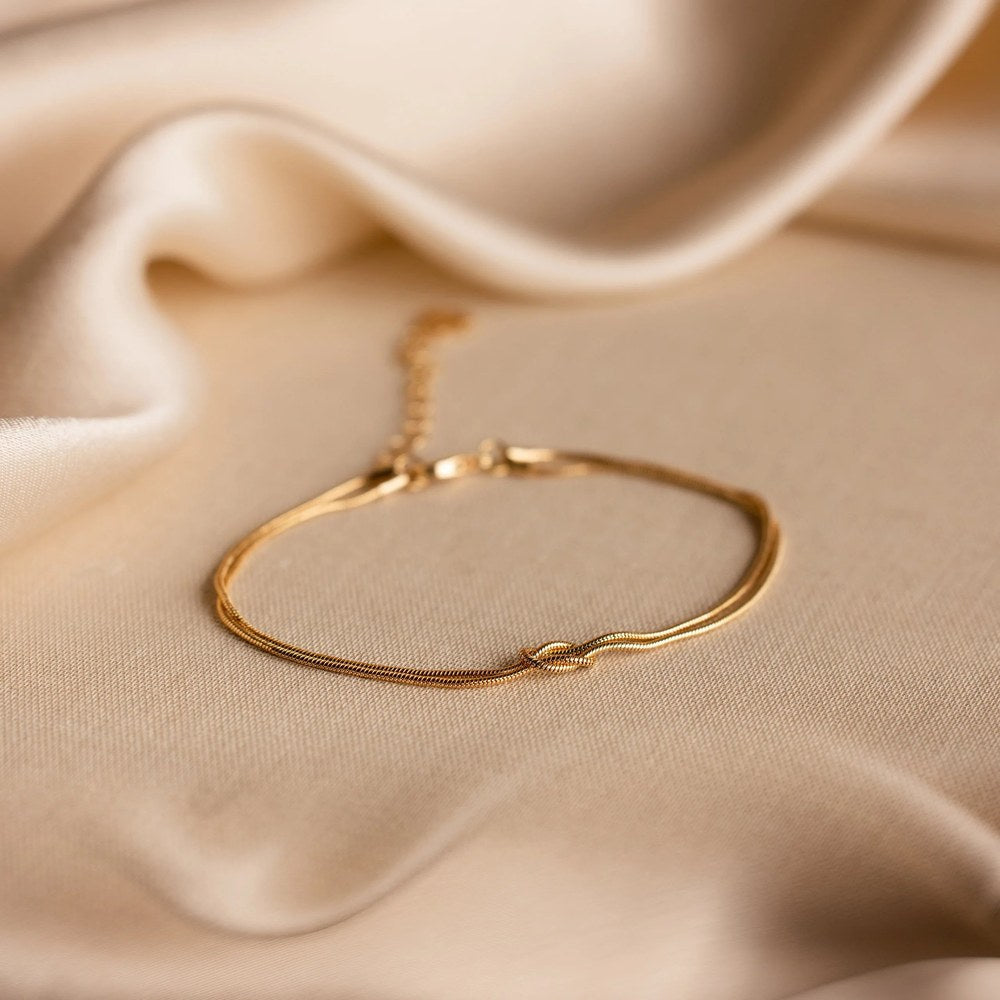 The Pomence Forever Knot Bracelet in warm 18k-gold tone photographed alone, clasped into a continuous ring on warm cream satin under directional light