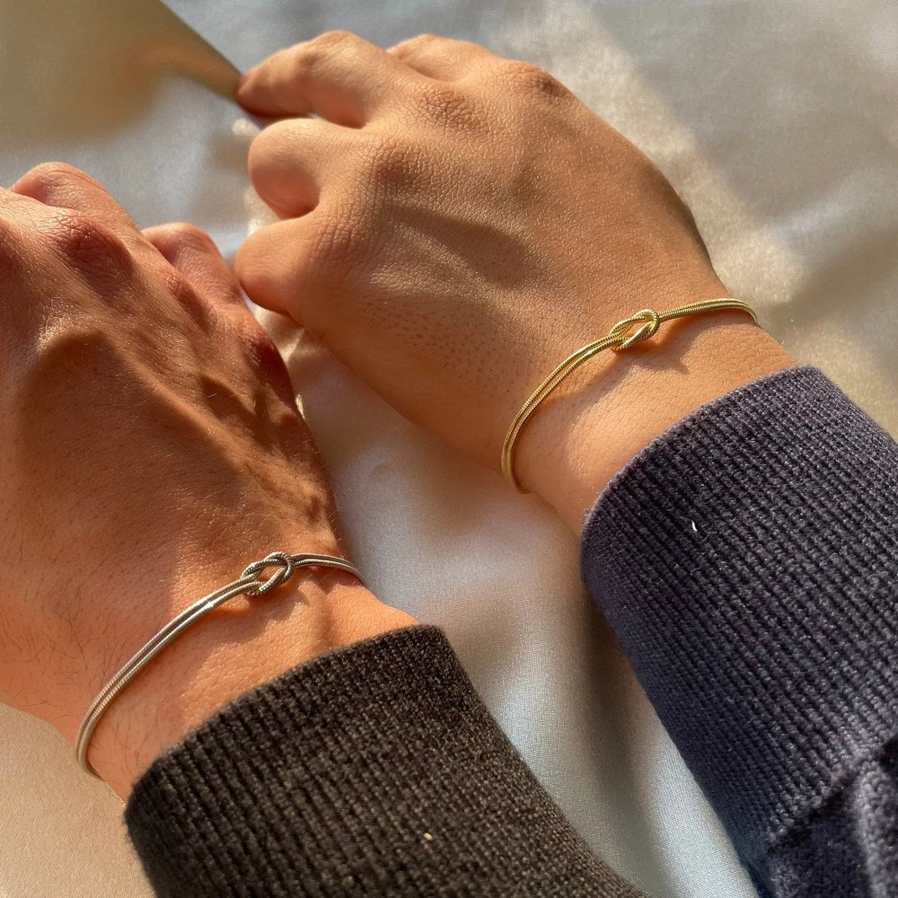 Two bare wrists together, one wearing the Pomence Forever Knot Bracelet in warm 18k-gold tone and one in cool steel-silver tone, both love knots reading