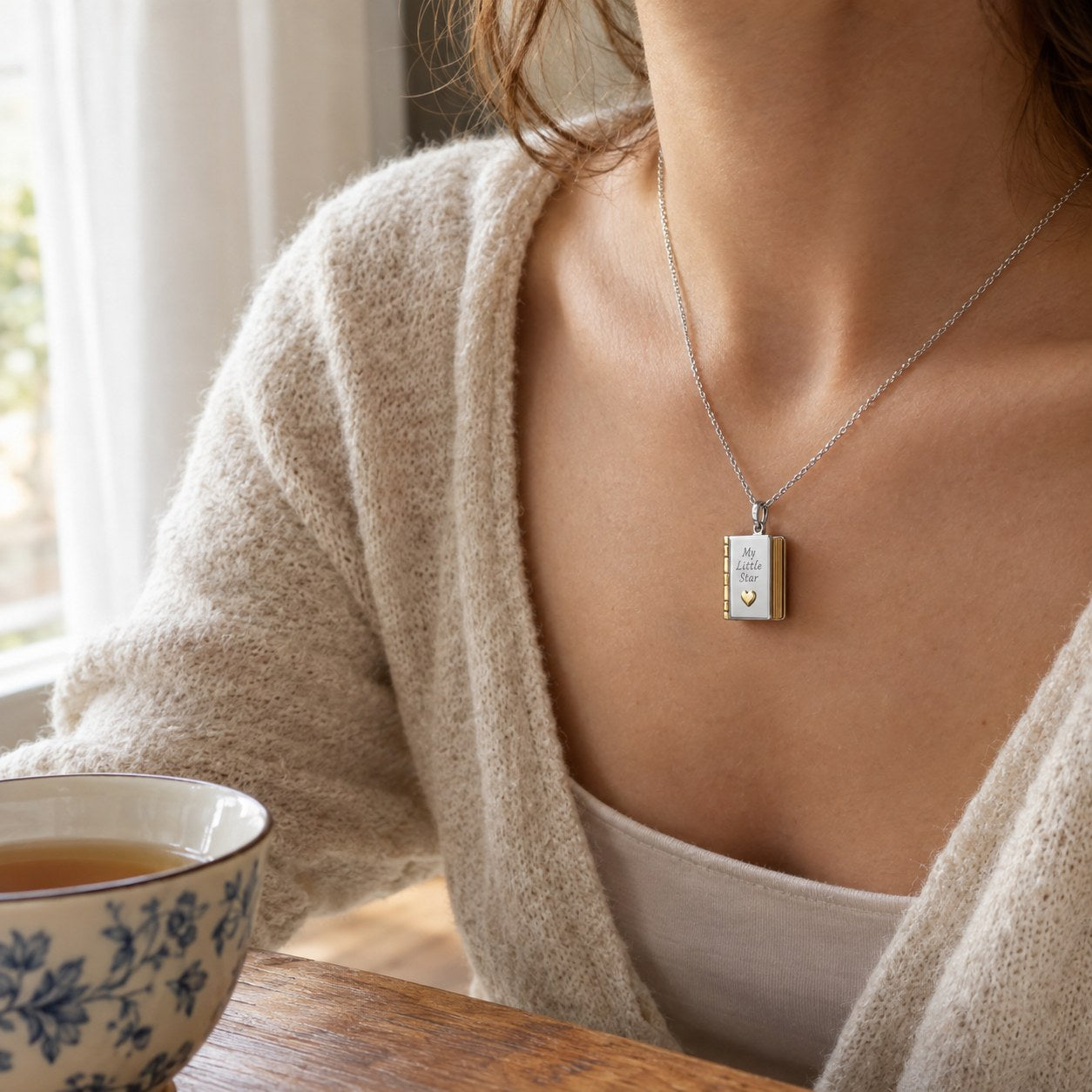 A woman at a table wearing the Pomence My Little Star Book Locket Necklace, the cover engraving visible at her collarbone