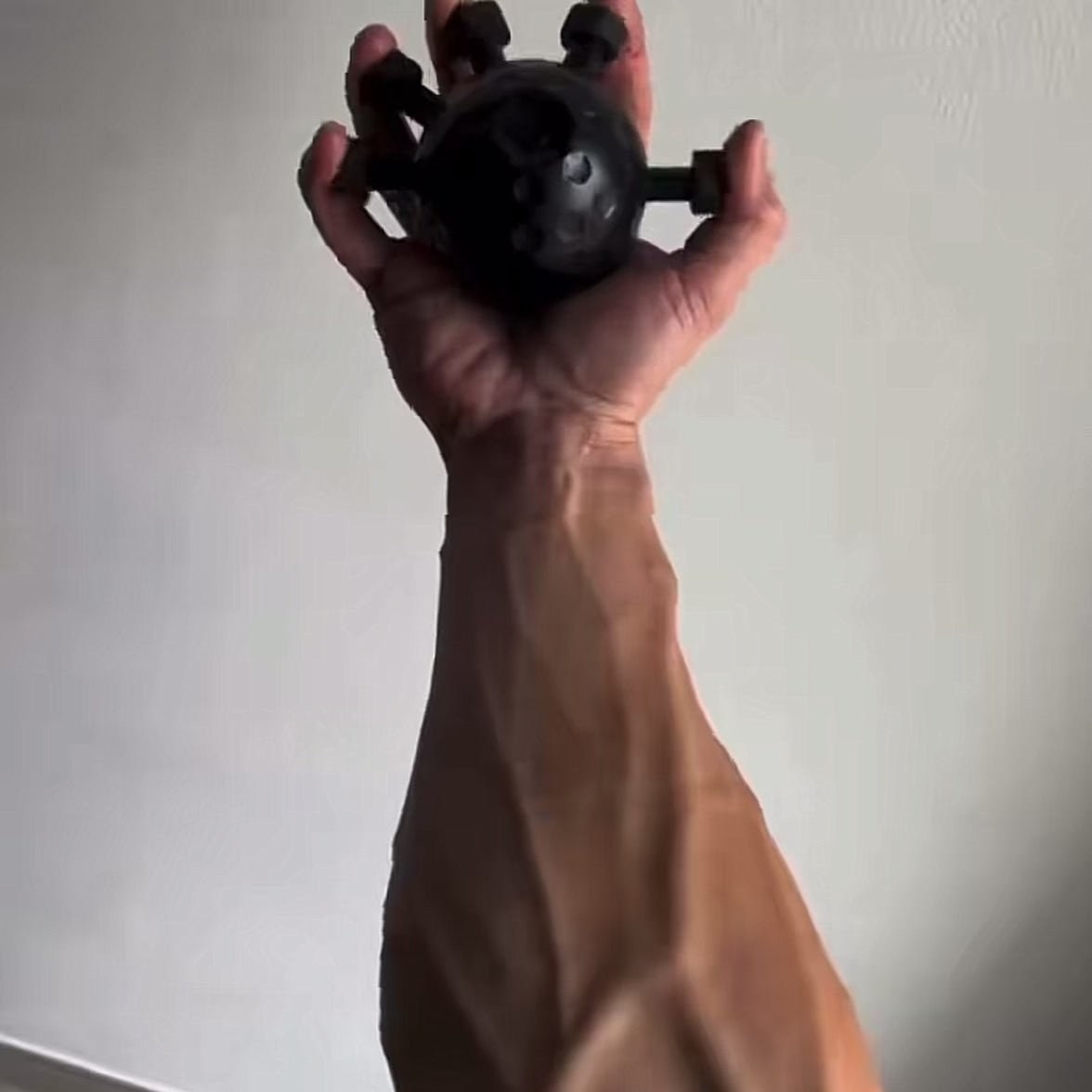 An adult hand squeezing the matte black Pomence Forearm Grip Trainer Ball with a fingertip hooked over each of the five pegs