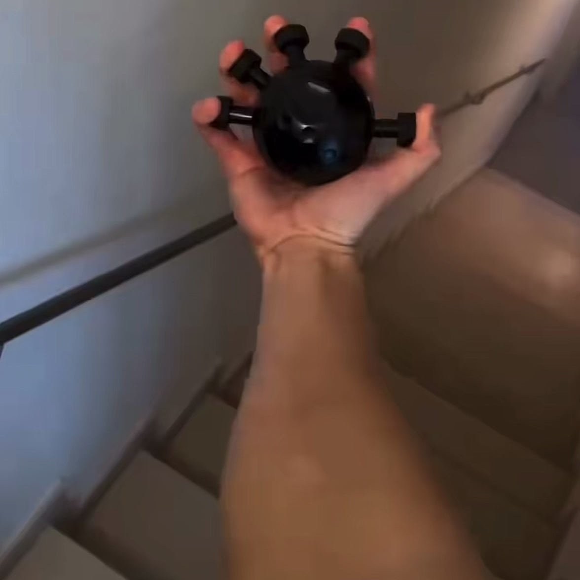 An adult hand spreading the five pegs of the matte black Pomence Forearm Grip Trainer Ball outward, forearm in frame