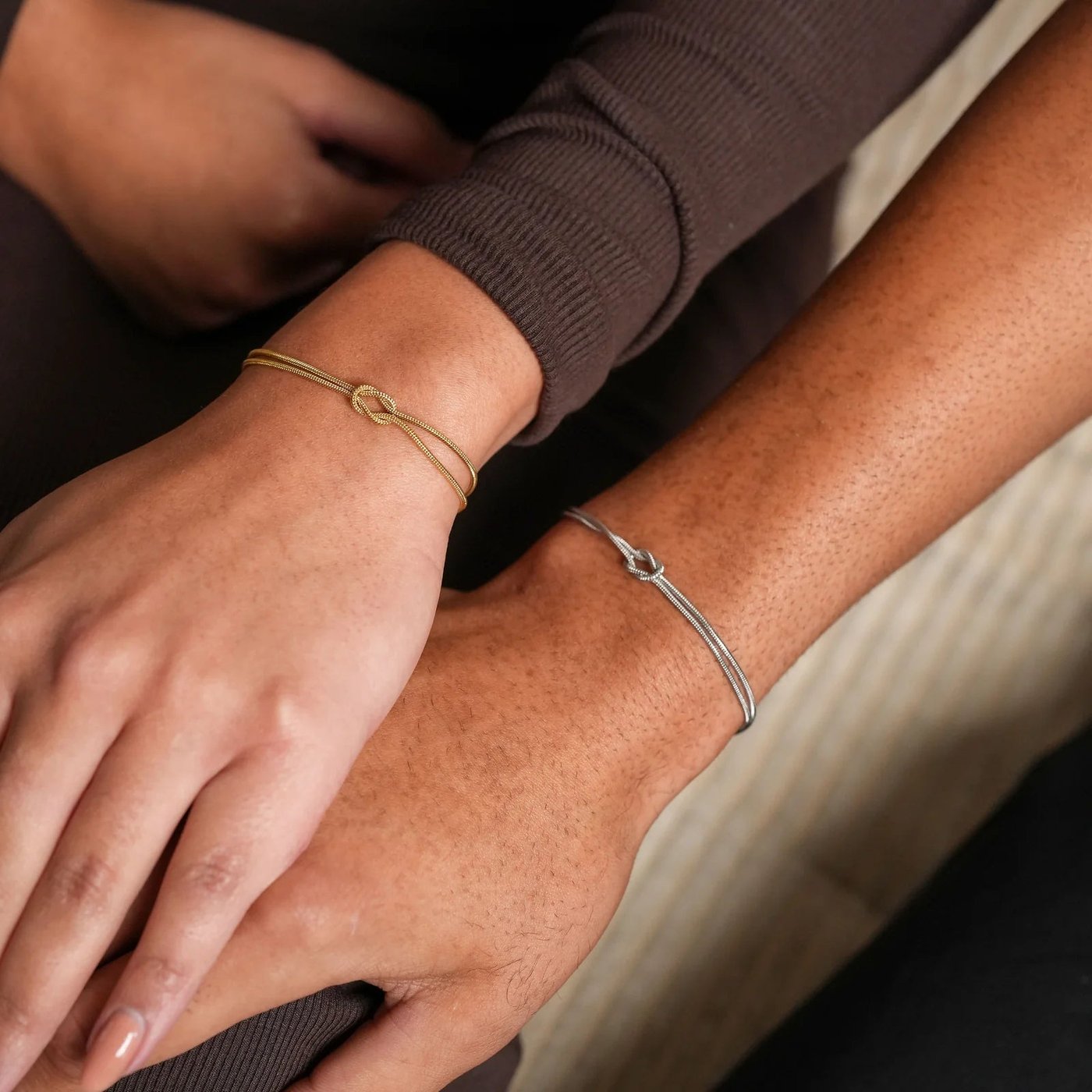 A man's wrist in cool steel-silver tone and a woman's wrist in warm 18k-gold tone side by side, both wearing the Pomence Forever Knot Bracelet