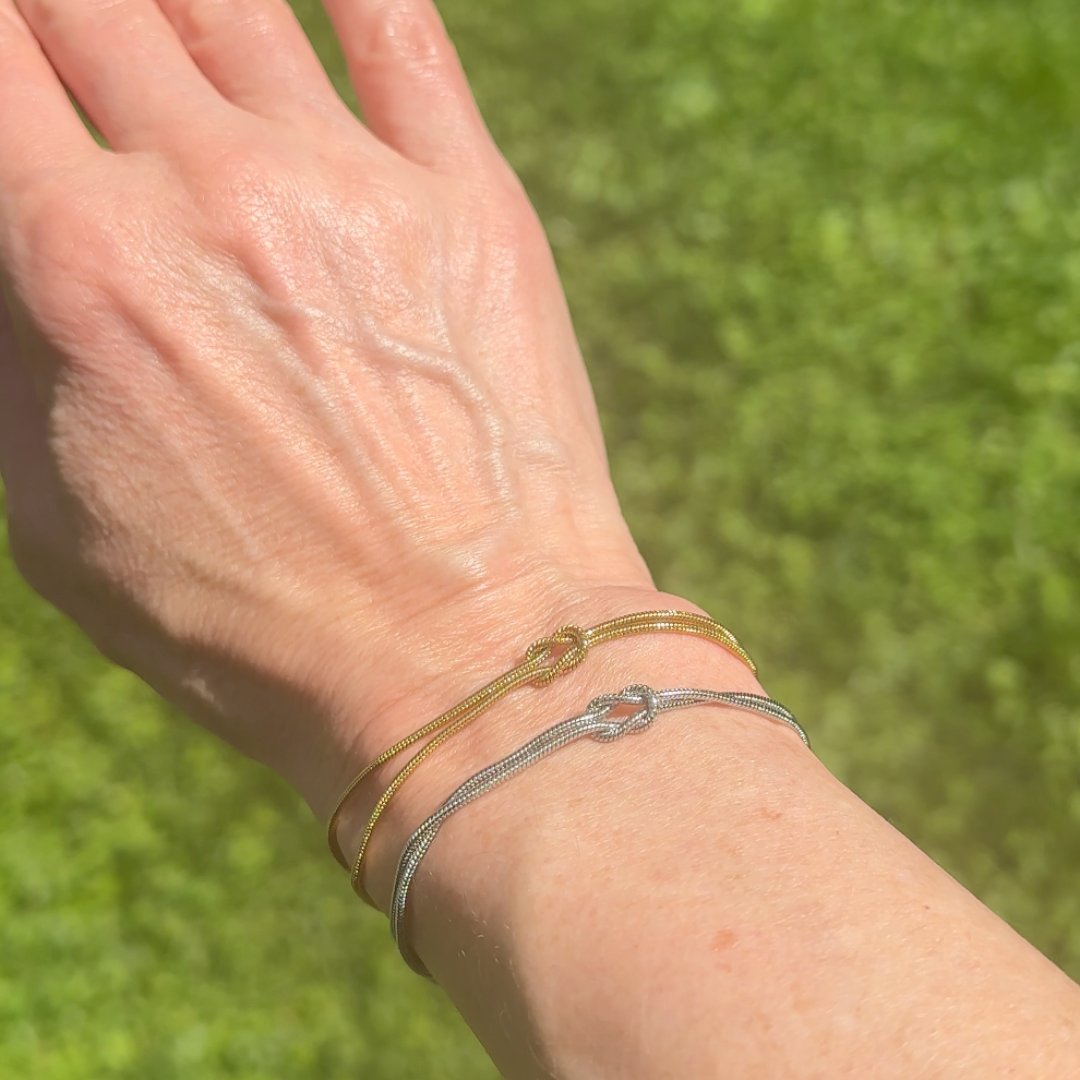 Water beaded on a bare wrist outdoors that is still wearing the Pomence Forever Knot Bracelet in warm 18k-gold tone and one in cool steel-silver tone