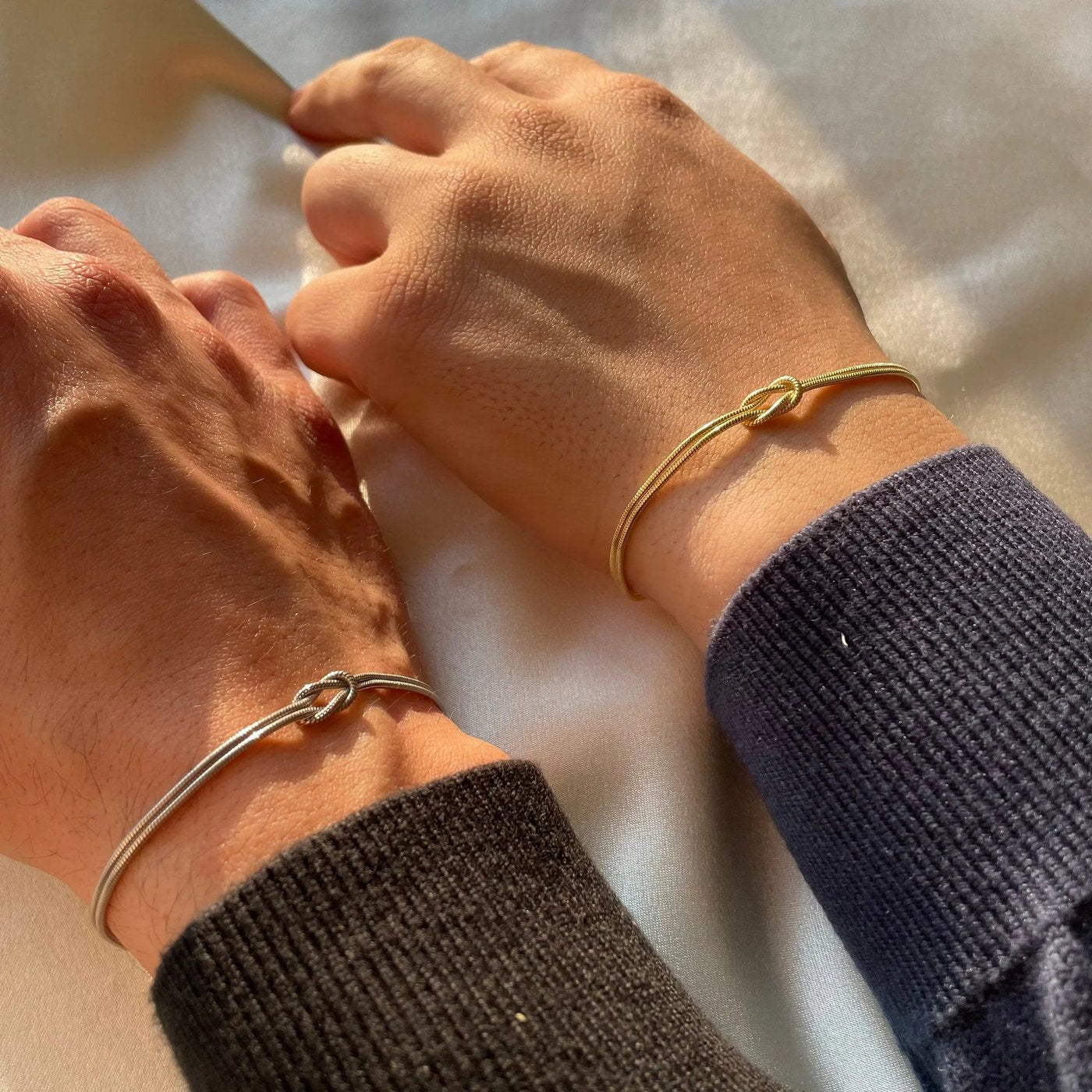 Two bare wrists resting together, one wearing the Pomence Forever Knot Bracelet in warm 18k-gold tone and one in cool steel-silver tone, the two knots side by side