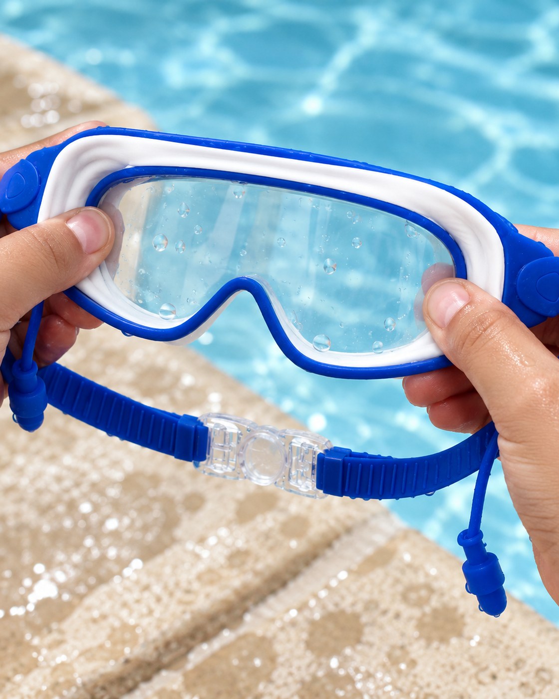 A hand pressing the clear clasp open on the strap of the Pomence Kids Swim Goggles at the edge of a pool