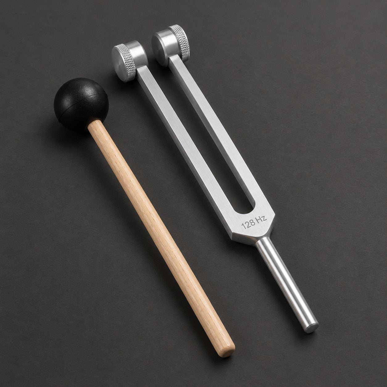 The Pomence 128 Hz Weighted Tuning Fork and its wooden mallet shown alone on a dark ground